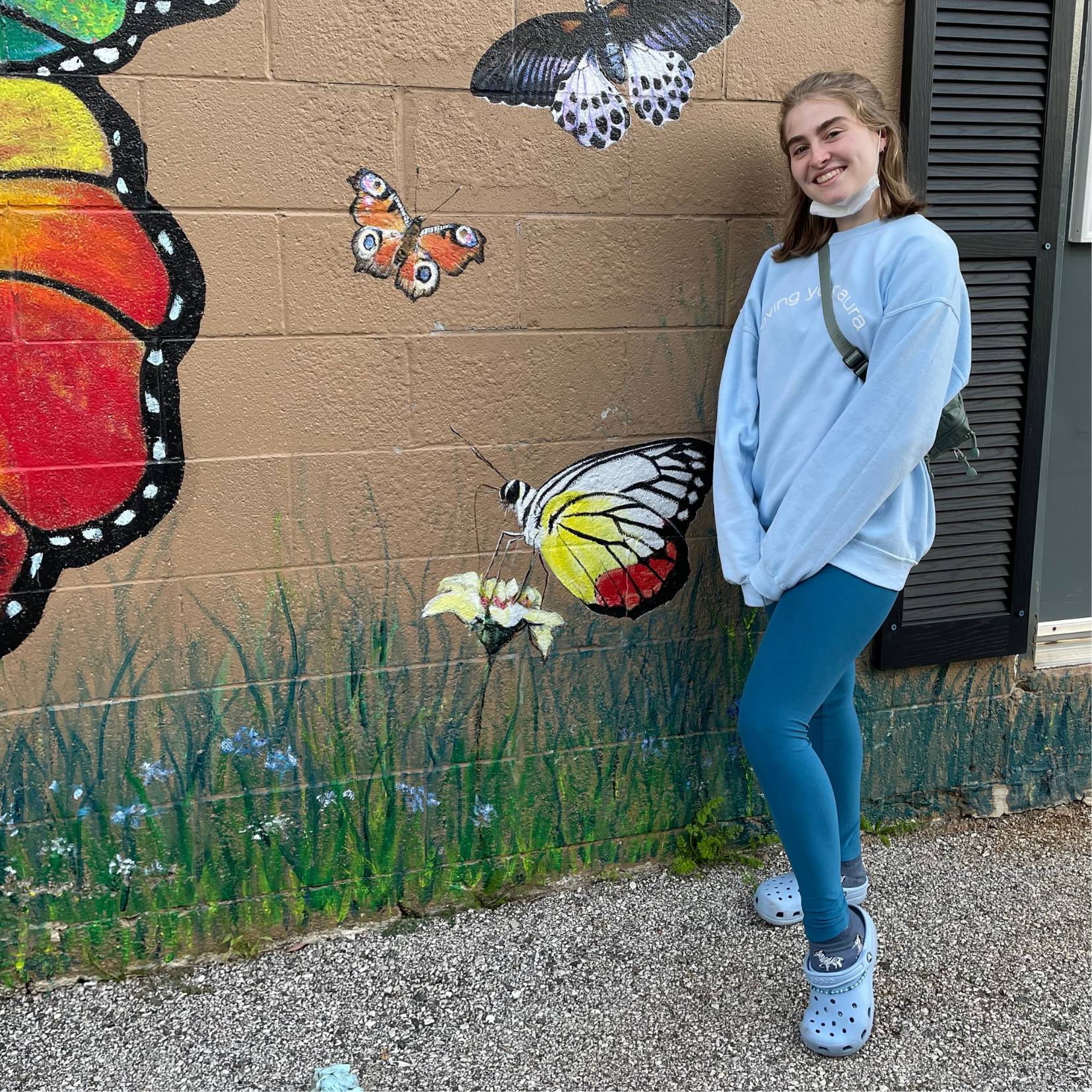 Image of female standing in front of a butterfly mural on a brick wall, smiling.